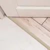 A close-up view of a floor transition between two different flooring materials, a marble-like tile and a wood-look plank, with a beige transition strip and a white baseboard above. Produktbild-Vorschau