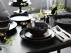 A table setting with dark stoneware plates, bowls, and a wine glass, decorated with green branches and lit candles. Náhľad obrázku produktu