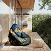 A bean bag chair with a colorful, abstract pattern is placed on a wooden deck next to a small woven stool. The scene is set outdoors, with lush greenery and a stone wall in the background, suggesting a relaxed, outdoor living space. Voir les images du produit