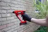 A person wearing a black glove is holding a red and black FERM cordless hammer drill against a brick wall. Produktbild-Vorschau