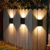 Three black solar-powered outdoor wall lights are mounted on a white wooden fence, illuminating the area above and below them. Náhľad obrázku produktu