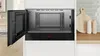An open built-in microwave oven is shown in a modern kitchen setting. The interior of the microwave is visible, along with its control panel. To the right, a wooden cutting board and two salt and pepper shakers are on a shelf. Voir les images du produit