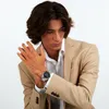 A man wearing a tan suit and white shirt, showing off a silver watch on his left wrist. Produktbild-Vorschau