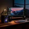 A computer desk setup at night with a large curved monitor displaying a mountain landscape, speakers, a keyboard, and a desk lamp. Produktbild-Vorschau