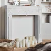A white, distressed wooden fireplace surround with decorative carvings, set against a plain wall. Several white pillar candles are arranged on the hearth in front of it, and a woven basket with a fluffy white item is to the left. Anteprima dell’immagine prodotto