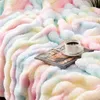 A soft, fluffy blanket with a rainbow pattern is draped over a surface, with a book and a cup of coffee placed on it. Produktbild-Vorschau