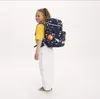 A young girl wearing a yellow t-shirt and white pants, with a colorful backpack on her back, is posing for a product photo against a white background. Voir les images du produit