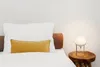 A close-up shot of a bed with white bedding and pillows, and a bedside table with a lamp. Produktbild-Vorschau