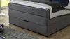 A close-up view of a grey upholstered box spring bed with a mattress and bedding, including a dark blue garment draped over the bed. Produktbild-Vorschau