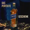 A bottle of Johnnie Walker Blue Label whisky is presented on a dark, textured surface with golden ribbons, suggesting a gift. Produktbild-Vorschau