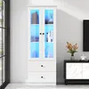 A white display cabinet with glass doors and LED lighting, showcasing decorative items and books. It is placed in a room with a window and a black cabinet with marble doors. Produktbild-Vorschau