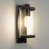 A modern black outdoor wall light with a clear glass cylinder and an exposed Edison-style bulb, mounted on a light gray wall. Produktbild-Vorschau