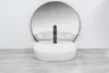A white oval countertop sink with a black faucet is placed on a wooden surface in front of a round mirror against a white wall. Náhľad obrázku produktu