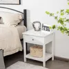A white nightstand with a drawer and an open shelf is placed next to a bed. Podgląd zdjęcia produktu