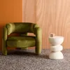 A green velvet armchair is placed next to a white side table with a mug on it, against a backdrop of orange and wood-paneled walls. Produktbild-Vorschau