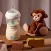 A baby bottle with milk and a plush monkey toy are displayed on a wooden surface. Produktbild-Vorschau
