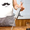 A person wearing an apron with a mustache print is holding a stainless steel meat injector over a wooden table. Voir les images du produit