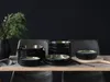 A set of black ceramic plates and bowls with a green and gold interior, arranged on a wooden table with a dark background and a vase of greenery. Náhľad obrázku produktu