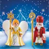 Two Playmobil figures, a St. Nicholas and a Christmas angel, stand in a snowy, festive scene with a church in the background. Produktbild-Vorschau
