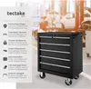 A black tool cart with drawers is shown in a workshop setting with blurred figures of people working in the background. Náhled obrázku produktu