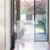 A white wooden pet gate is set up in a doorway, leading to a backyard with lush greenery and trees. Podgląd zdjęcia produktu