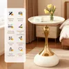 A round white side table with a gold pedestal base, placed next to a bed and nightstand in a bedroom setting. The table has a vase of yellow tulips and a small white object on top. Produktbild-Vorschau
