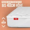 A close-up of a white mattress topper with a red brand tag, set against an orange, wavy background. Produktbild-Vorschau