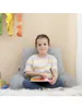 A young girl sits on a fluffy rug, leaning against a large gray pillow, playing with a wooden toy. Anteprima dell’immagine prodotto