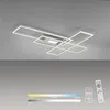 A modern LED ceiling light fixture with a geometric design, shown mounted on a gray ceiling. Below the fixture, there is a color temperature and brightness scale, along with a remote control. Produktbild-Vorschau
