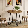 A round wooden dining table with black metal legs, set in a room with two chairs and framed posters on the wall. Produktbild-Vorschau