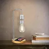 A levitating LED light bulb sits above a wooden base with a smartphone on it, placed on a wooden table next to a stack of books. Náhled obrázku produktu