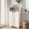 A white bathroom cabinet with open shelves, a drawer, and two doors, placed in a bathroom setting with tiled walls and a window. Náhled obrázku produktu