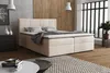 A bedroom scene featuring a beige boxspring bed with storage, two pillows, and a draped blanket. The room has wooden walls, a large window with blinds, and a patterned rug. Voir les images du produit