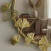 A close-up of a gold-colored decorative mirror frame with ginkgo leaf motifs, with a blurred background showing a wooden table and chair. Voir les images du produit