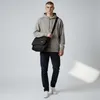A man wearing a grey hoodie and dark jeans stands against a white background, holding a black messenger bag. Produktbild-Vorschau
