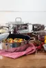 A collection of stainless steel pots and pans, some filled with food, arranged on a wooden surface with a red and white checkered cloth and tomatoes. Náhled obrázku produktu