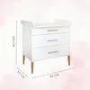 A white changing table with three drawers and wooden legs, shown against a soft pink watercolor background. Dimensions are indicated with lines and text. Podgląd zdjęcia produktu