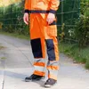 A person wearing orange work pants with navy blue accents and reflective stripes stands on a paved walkway next to a grassy area with a fence in the background. Anteprima dell’immagine prodotto