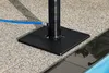 A close-up view of a black base and pole for a garden shower, with a blue hose connected to it, situated on a speckled patio next to a pool. Produktbild-Vorschau