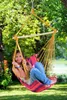 A young woman is relaxing in a colorful hammock chair outdoors, surrounded by trees and grass. Produktbild-Vorschau