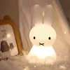A cute bunny-shaped night light is placed on a fluffy white rug, next to a framed picture. The scene is illuminated by fairy lights, creating a cozy atmosphere. Produktbild-Vorschau