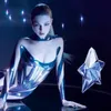 A woman in a futuristic silver outfit poses next to a star-shaped perfume bottle. Voir les images du produit