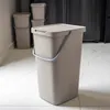 A brown plastic trash can with a lid and handle is shown in a kitchen setting, with other similar trash cans in the background. Podgląd zdjęcia produktu