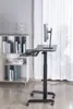 A black adjustable standing desk with a monitor, keyboard, and mouse is shown in a room with a bookshelf in the background. Voir les images du produit