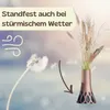 A vase with dried flowers is shown, with a dandelion being blown by wind to illustrate stability in windy conditions. Produktbild-Vorschau