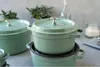 Several green Staub cast iron pots are stacked and arranged on a gray surface, with some greenery and a glass bottle in the background. Produktbild-Vorschau