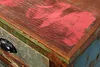 A close-up view of the distressed wooden surface of a piece of furniture, showing painted accents and a drawer pull. Náhled obrázku produktu