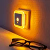 A night light plugged into a wall outlet, casting a warm amber glow on the wall and a pair of glasses on a surface in the foreground. Produktbild-Vorschau