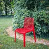 A red chair sits on a lawn in front of a dense green bush. Produktbild-Vorschau