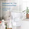 A BRITA water filter pitcher is shown on a table with two glass bottles in the foreground. The background is blurred, showing a plant and window. Produktbild-Vorschau